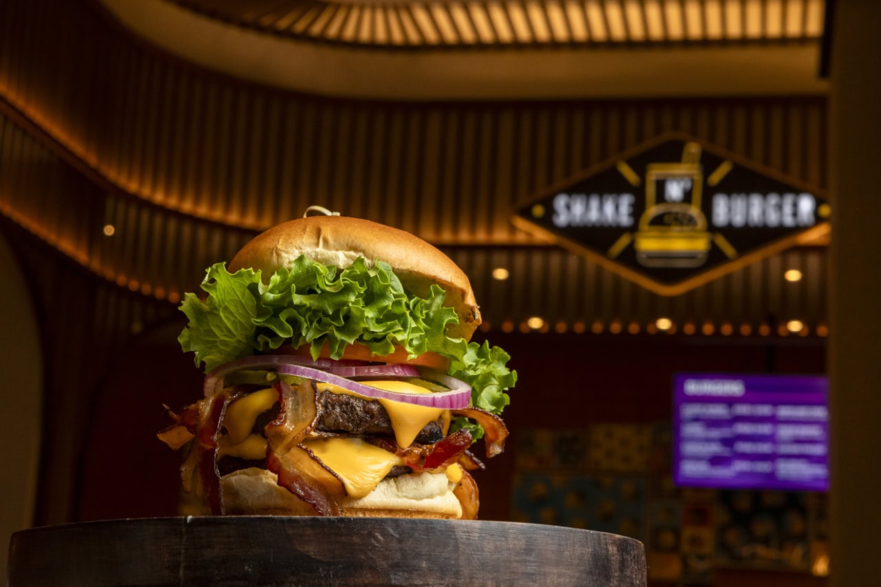 A large burger placed on a wooden stand, showcasing its layers of ingredients and toasted bun.