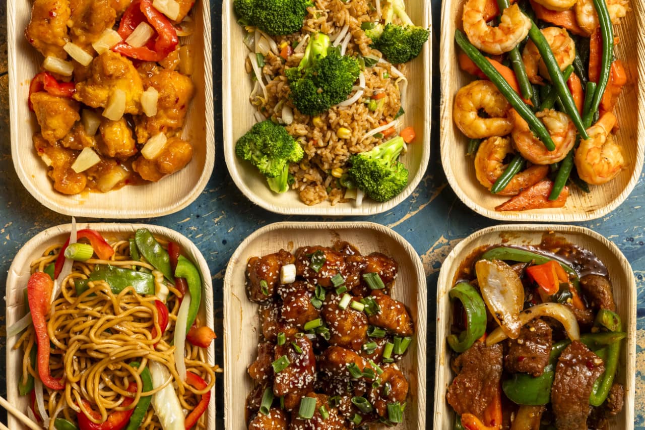 A selection of Asian dishes in plastic containers, noodles, and stir-fried vegetables, arranged on a table.